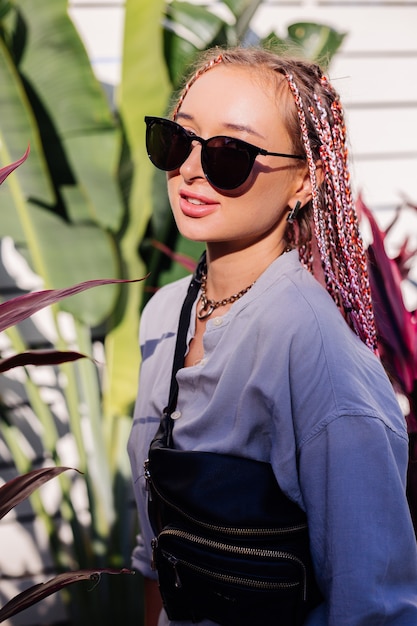 Foto grátis jovem mulher elegante com tranças rosa roxas e bolsa preta na cintura posando ao ar livre