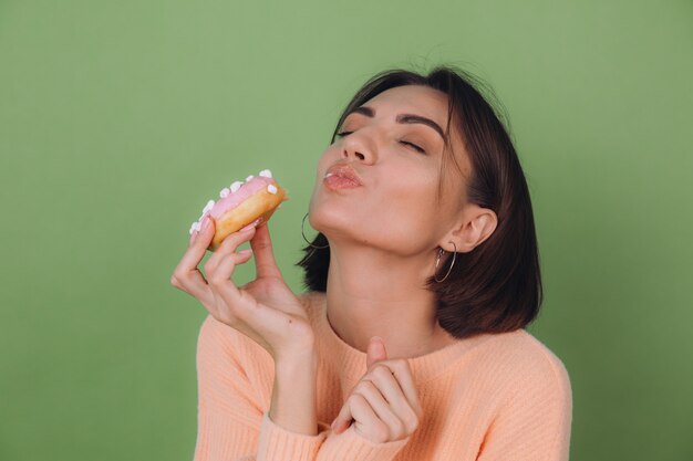 Jovem mulher elegante com um suéter casual cor de pêssego e óculos laranja, isolado na parede verde oliva, mordendo um donut rosa com os olhos fechados copiar o espaço