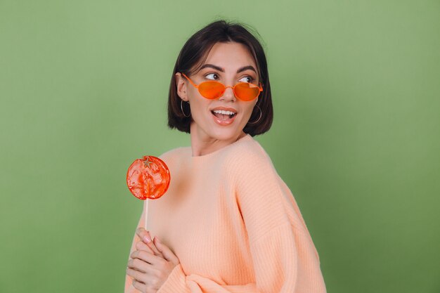 Jovem mulher elegante com um suéter casual cor de pêssego e óculos laranja isolados em uma parede verde oliva com um pirulito laranja sorriso positivo e espaço de cópia