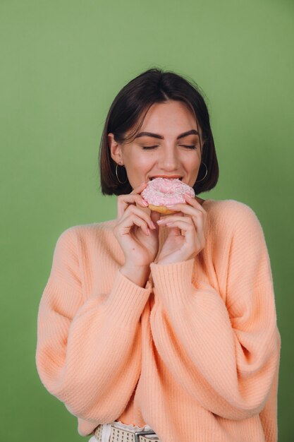 Jovem mulher elegante com um suéter casual cor de pêssego isolado em uma parede verde-oliva com rosquinha rosa e espaço para cópia