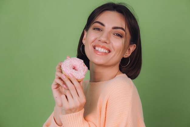 Jovem mulher elegante com um suéter casual cor de pêssego isolado em uma parede verde-oliva com rosquinha rosa e espaço para cópia