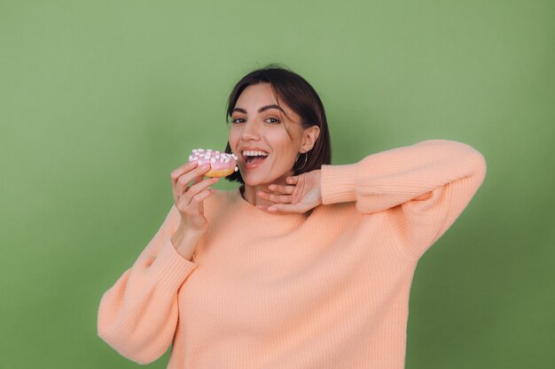 Jovem mulher elegante com um suéter casual cor de pêssego isolado em uma parede verde-oliva com rosquinha rosa e espaço para cópia