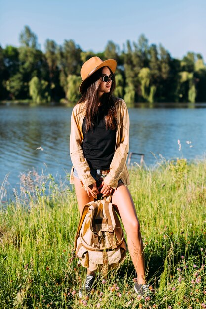 Jovem mulher elegante em pé perto do rio com mochila de exploração