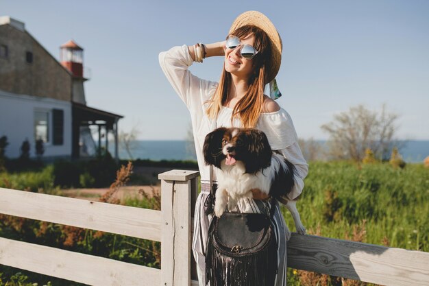 Jovem mulher elegante no campo, segurando um cachorro, feliz humor positivo, verão, chapéu de palha, roupa de estilo boêmio, óculos de sol, sorrindo, feliz, ensolarado