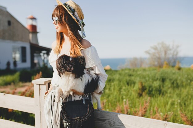 Jovem mulher elegante no campo, segurando um cachorro, feliz humor positivo, verão, chapéu de palha, roupa de estilo boêmio, óculos de sol, sorrindo, feliz, ensolarado