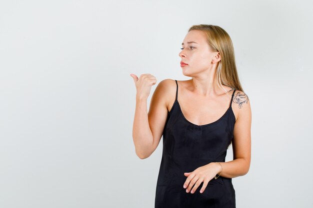 Jovem mulher em camiseta preta apontando para trás com o polegar e olhando séria