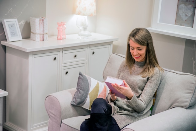 Jovem mulher em casa sentada na cadeira moderna relaxando no livro de leitura da sala de estar. Livro de leitura de jovem mulher, no interior da casa