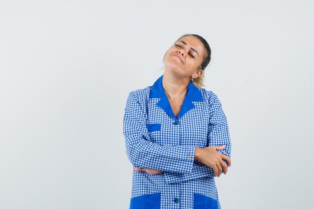 Jovem mulher em pé com os braços cruzados na camisa de pijama azul guingão e olhando calma, vista frontal.