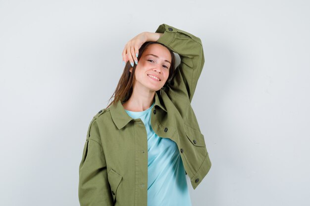 Jovem mulher em t-shirt, jaqueta posando com a mão levantada na cabeça e olhando alegre, vista frontal.