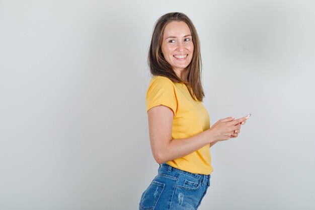 Jovem mulher em t-shirt, shorts, olhando para longe com o smartphone nas mãos e olhando alegre.