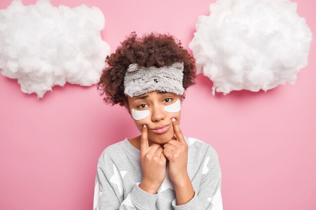 Foto grátis jovem mulher encaracolada triste força o sorriso mantém os dedos perto dos cantos dos lábios olha tristemente para a câmera usa pijama de máscara de dormir tem uma expressão sonolenta quando acorda cedo de manhã fica em casa fica chateado