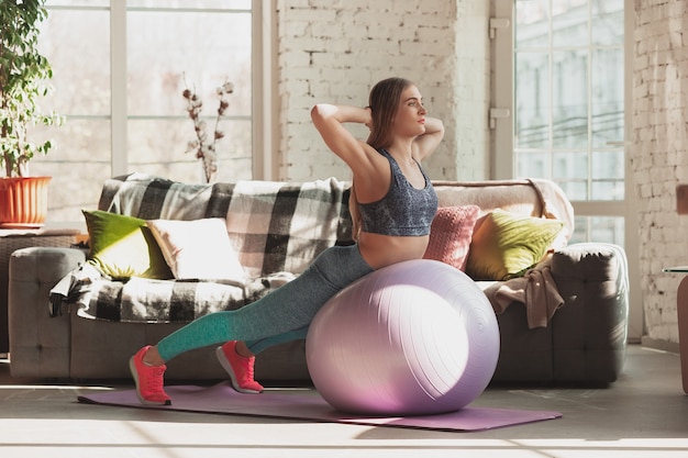 Jovem mulher ensinando em casa cursos on-line de fitness, estilo de vida aeróbio e esportivo durante a quarentena. Ficar ativo enquanto isolado, bem-estar, conceito de movimento. Exercícios com fitball para a parte inferior do corpo.