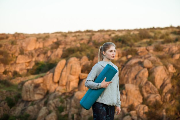 Jovem mulher esportiva segurando o tapete de ioga, em pé na pedra no canyon