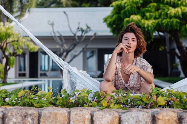 Foto grátis jovem mulher europeia fumando charuto deitada na rede em frente a um luxuoso hotel villa tropical, luz natural ao pôr do sol