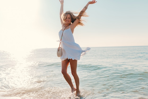 Jovem mulher feliz dançando virando à beira-mar praia ensolarada verão estilo da moda vestido branco férias