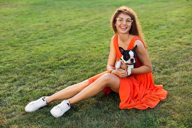 Jovem mulher feliz e sorridente com vestido laranja se divertindo brincando com o cachorro no parque, estilo verão, clima alegre