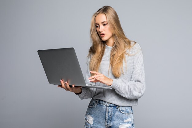 Jovem mulher feliz e sorridente em roupas casuais segurando um laptop e enviando e-mail para a melhor amiga, isolada em um fundo cinza
