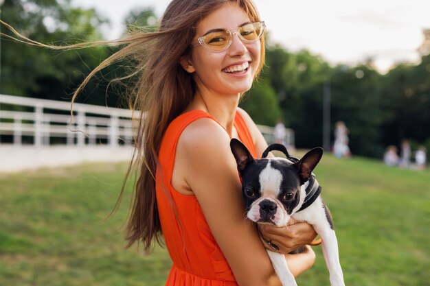 Jovem mulher feliz e sorridente segurando cachorro Boston Terrrier no parque, dia ensolarado de verão, clima alegre, brincar com o animal de estimação, acenar cabelo comprido, se divertir, tendência da moda para o verão