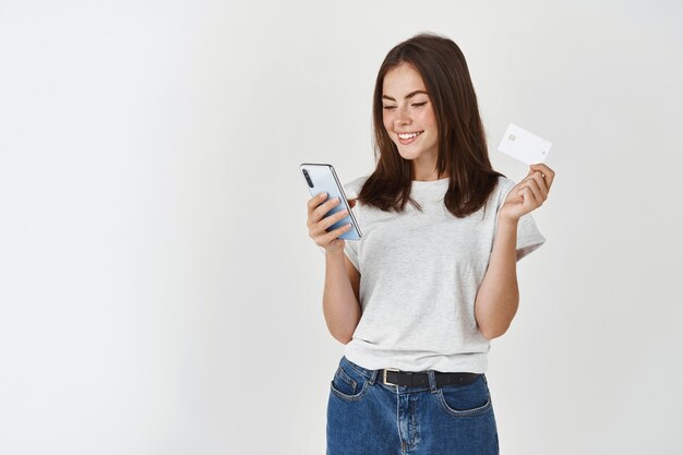 Jovem mulher feliz, pagando com smartphone e cartão de crédito de plástico, em pé sobre uma parede branca.