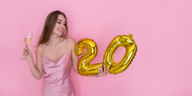 Jovem mulher feliz segurando um balão de folha de ouro e taça de champanhe na festa de aniversário de fundo rosa
