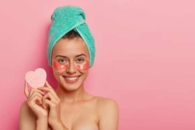 Jovem mulher feliz usa adesivos hidratantes de colágeno sob os olhos, segura uma esponja para remover a maquiagem, faz tratamentos de beleza, fica com a pele fresca após o banho