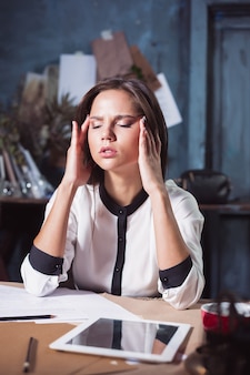 Jovem mulher frustrada trabalhando em um loft para casa ou em uma mesa de escritório em frente a um laptop sofrendo de dores de cabeça crônicas diárias