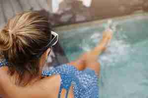 Foto grátis jovem mulher grávida com nó de cabelo e pele bronzeada, sentada na beira da piscina e espirrando água nas pernas.