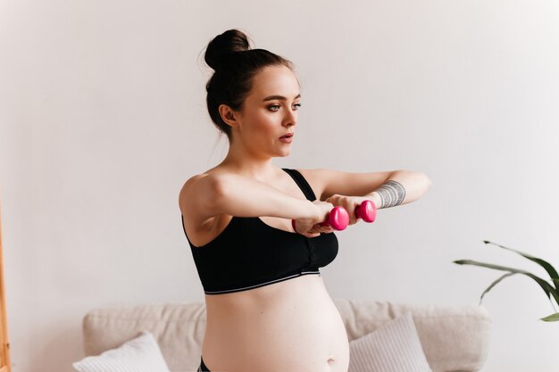 Jovem mulher grávida em preto recortado top detém halteres rosa. Menina morena fazendo esporte na acolhedora sala de estar leve.