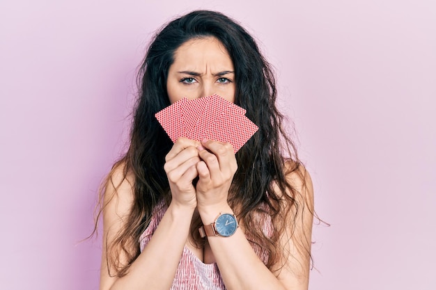 Jovem mulher hispânica cobrindo a boca com cartas céticas e nervosas franzindo a testa chateadas por causa de um problema de pessoa negativa