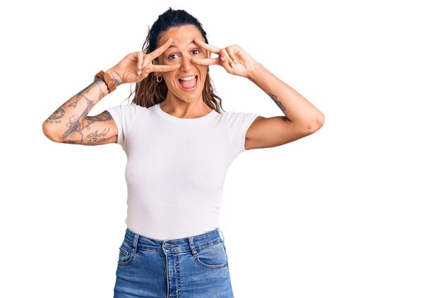 Jovem mulher hispânica com tatuagem vestindo camiseta branca casual fazendo o símbolo da paz com os dedos sobre o rosto, sorrindo alegre mostrando vitória