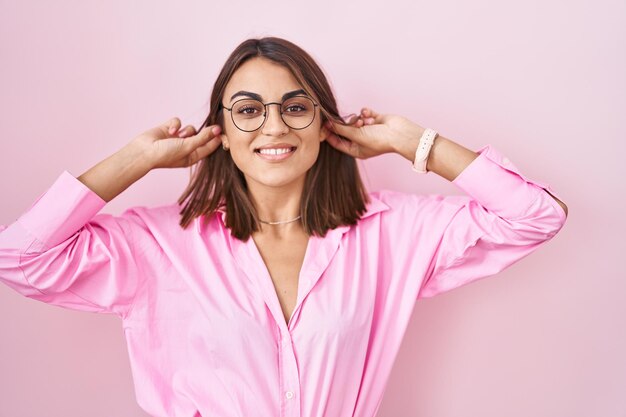 Jovem mulher hispânica de óculos em cima de fundo rosa sorrindo puxando as orelhas com os dedos, gesto engraçado. problema de audição