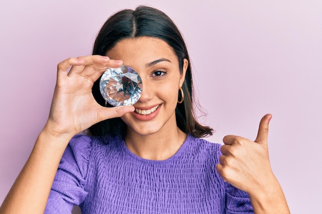 Jovem mulher hispânica segurando pedra de diamante brilhante sorrindo feliz e positivo, polegar para cima fazendo excelente e sinal de aprovação