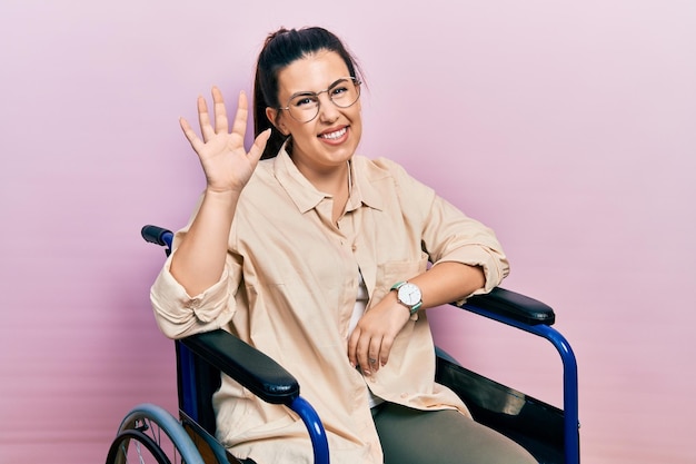 Jovem mulher hispânica sentada na cadeira de rodas, dizendo olá feliz e sorridente gesto de boas-vindas amigável
