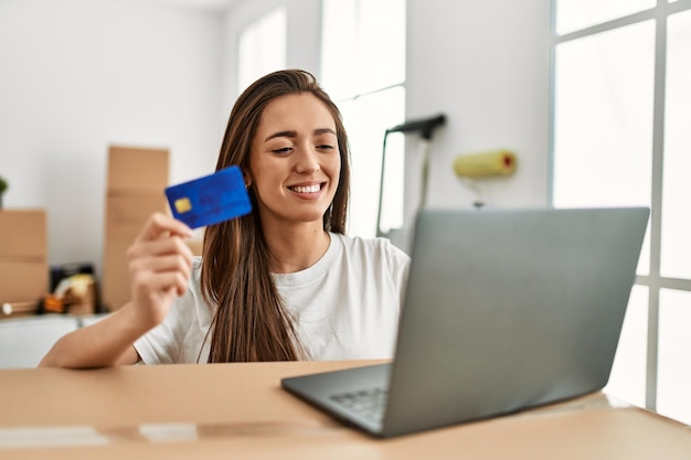 Jovem mulher hispânica sorrindo confiante usando cartão de crédito e laptop em nova casa