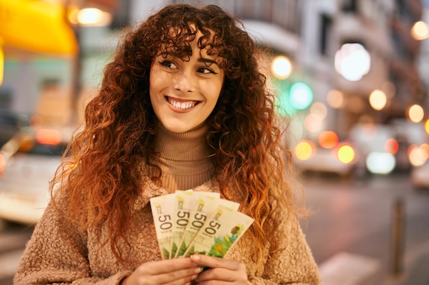 Jovem mulher hispânica sorrindo feliz segurando notas de israel shekels na cidade