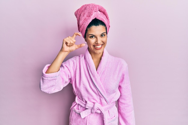 Jovem mulher hispânica usando touca de banho e roupão de banho sorrindo e confiante gesticulando com a mão fazendo sinal de tamanho pequeno com os dedos olhando e o conceito de medida da câmera
