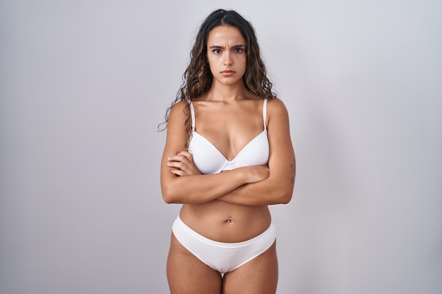 Jovem mulher hispânica vestindo lingerie branca cética e nervosa, desaprovando a expressão no rosto com os braços cruzados. pessoa negativa.