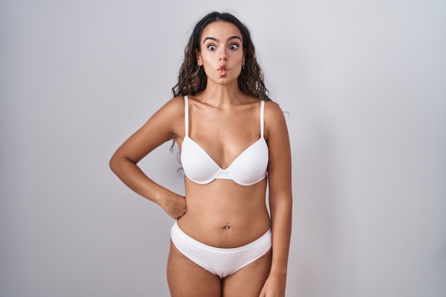 Jovem mulher hispânica vestindo lingerie branca fazendo cara de peixe com os lábios, gesto louco e cômico. expressão engraçada.