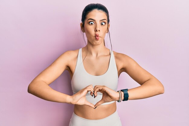 Jovem mulher hispânica vestindo roupas esportivas fazendo o símbolo do coração com as mãos fazendo cara de peixe com a boca e os olhos semicerrados, loucos e cômicos.