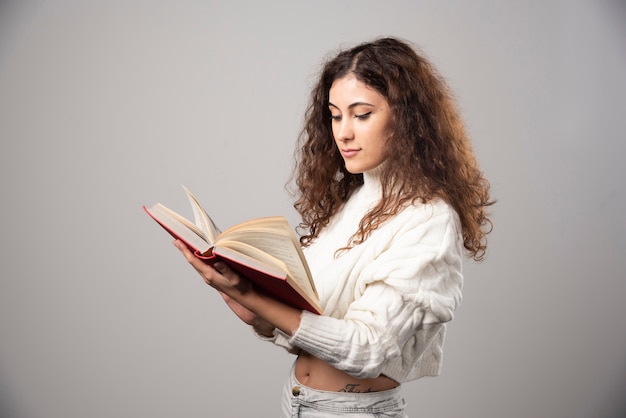 Foto grátis jovem mulher lendo um livro vermelho em uma parede cinza. foto de alta qualidade