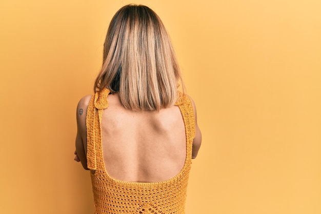 Jovem mulher loira caucasiana vestindo camiseta amarela casual em pé para trás olhando para longe com os braços cruzados