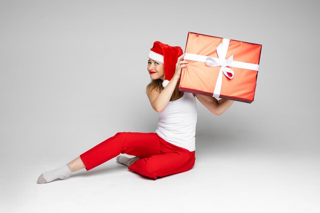 Jovem mulher loira com chapéu de Papai Noel parecendo perturbada, segurando uma grande caixa vermelha com presentes de Natal. Conceito de férias