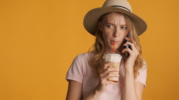 Jovem mulher loira de chapéu bebendo café enquanto conversa séria ao telefone sobre fundo colorido. Copiar espaço