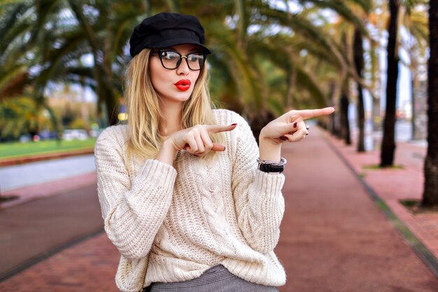 Jovem mulher loira elegante mostrando a direção por seus dedos, saiu de emoções surpresas, roupa de glamour elegante na moda, ruas de Barcelona com palmas, clima de viagem.