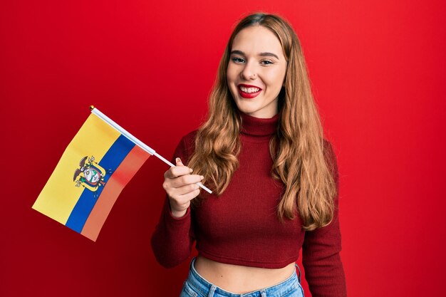 Jovem mulher loira segurando a bandeira do equador parecendo positiva e feliz de pé e sorrindo com um sorriso confiante mostrando os dentes