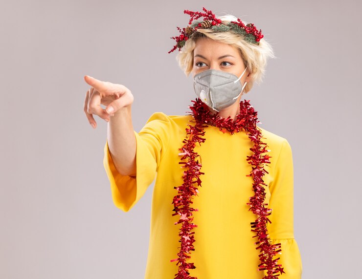 Foto grátis jovem mulher loira usando coroa de natal e guirlanda de ouropel em volta do pescoço com máscara protetora olhando e apontando para o lado isolado no fundo branco