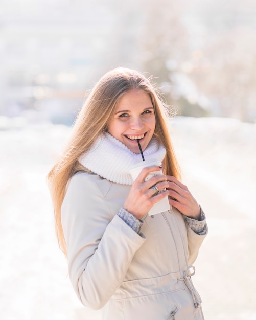 Jovem mulher loura de sorriso que bebe o copo de café descartável no inverno