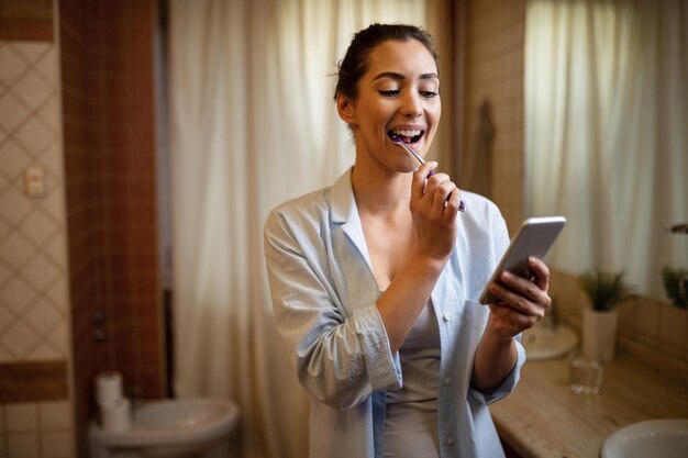 Jovem mulher mandando mensagens no celular enquanto escova os dentes no banheiro