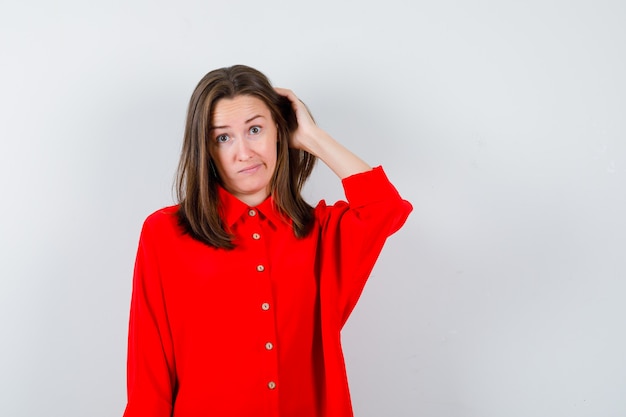 Jovem mulher mantendo a mão na cabeça com blusa vermelha e olhando hesitativa, vista frontal.