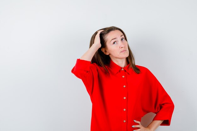 Jovem mulher mantendo a mão na cabeça, olhando para cima com uma blusa vermelha e parecendo pensativa. vista frontal.